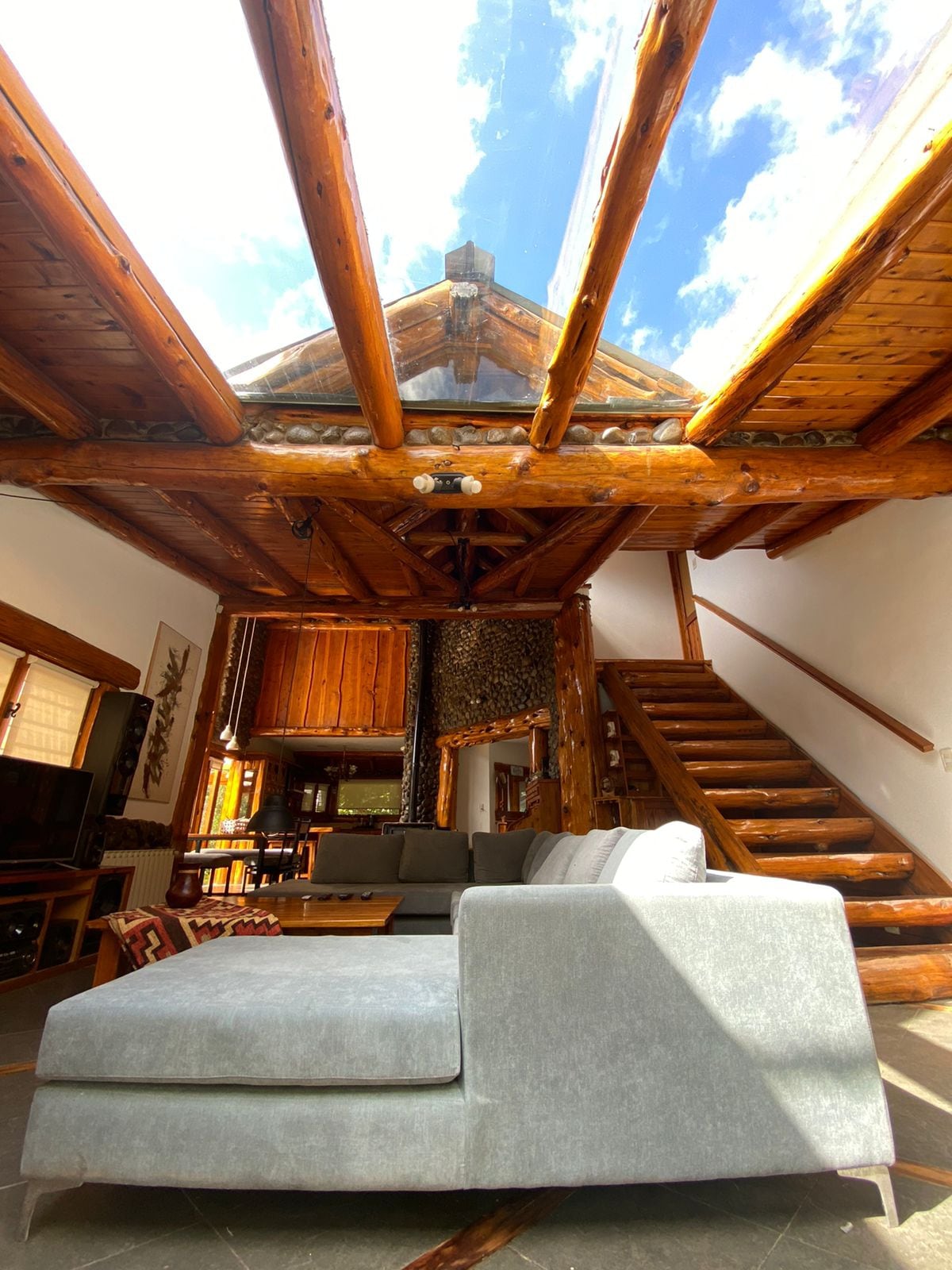 A spacious living area features large glass panels in the ceiling, allowing natural light to fill the room. Wooden beams and walls create an inviting texture, complemented by a modern sofa. A staircase leads to the upper level, enhancing the open feel of the space.