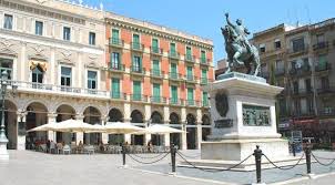 An urban plaza is depicted, featuring a prominent equestrian statue on a stone pedestal at the center. Surrounding the statue, outdoor seating areas with canopies are visible. Historic buildings with balconies and colorful facades create a lively atmosphere, complemented by clear blue skies.