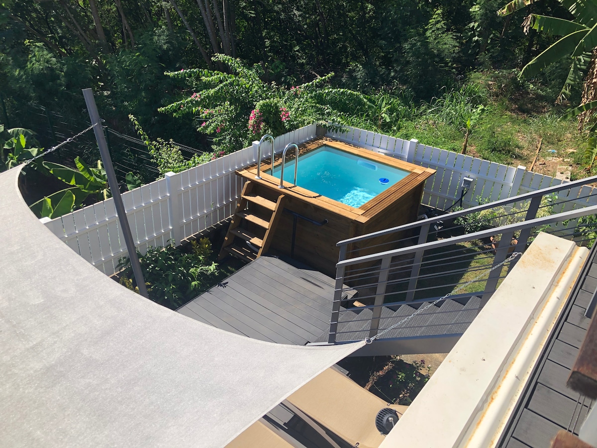 An elevated view showcases a wooden hot tub surrounded by lush greenery. Steps lead down from a deck to the hot tub area, enclosed by a white fence. Sunlight reflects off the clear water, contributing to a serene outdoor setting.