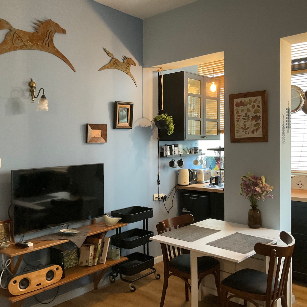 A cozy corner of the apartment features a small white dining table with two chairs. A television is positioned on an open shelf beside decorative wall art. The kitchen is visible through an archway, showcasing cabinetry and kitchen appliances. Natural light filters in through partially open blinds.