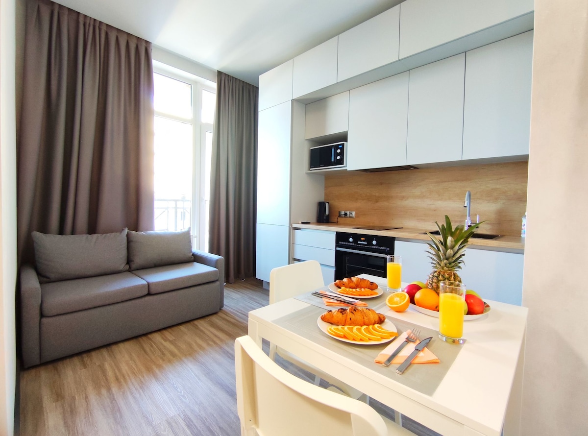 A modern kitchen area is featured, showcasing a light wooden countertop with a breakfast table set for two. A gray sofa is visible alongside large windows draped with dark curtains, allowing natural light to brighten the space. Fresh fruit and orange juice are displayed on the table.