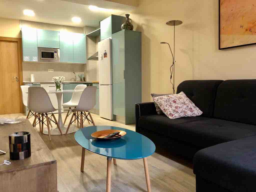 A modern living area features a dark sofa accompanied by a small round coffee table with a blue surface. A dining table with white chairs is situated nearby, while contemporary light fixtures and a kitchenette with mint green cabinetry are visible in the background.