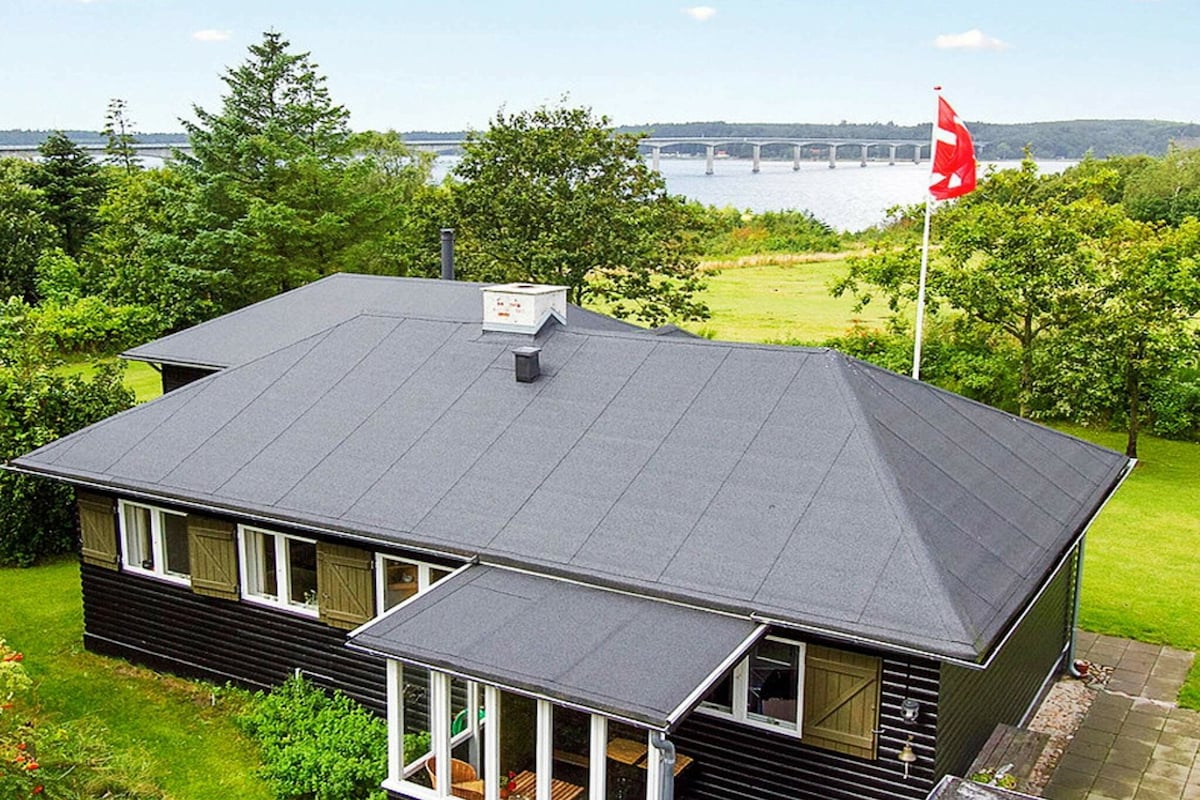 The exterior of the cottage showcases a black wooden structure with a gray roof. Large windows provide views of lush greenery and the nearby Limfjord, while a Danish flag flutters in the breeze. A covered terrace extends from the main entrance.