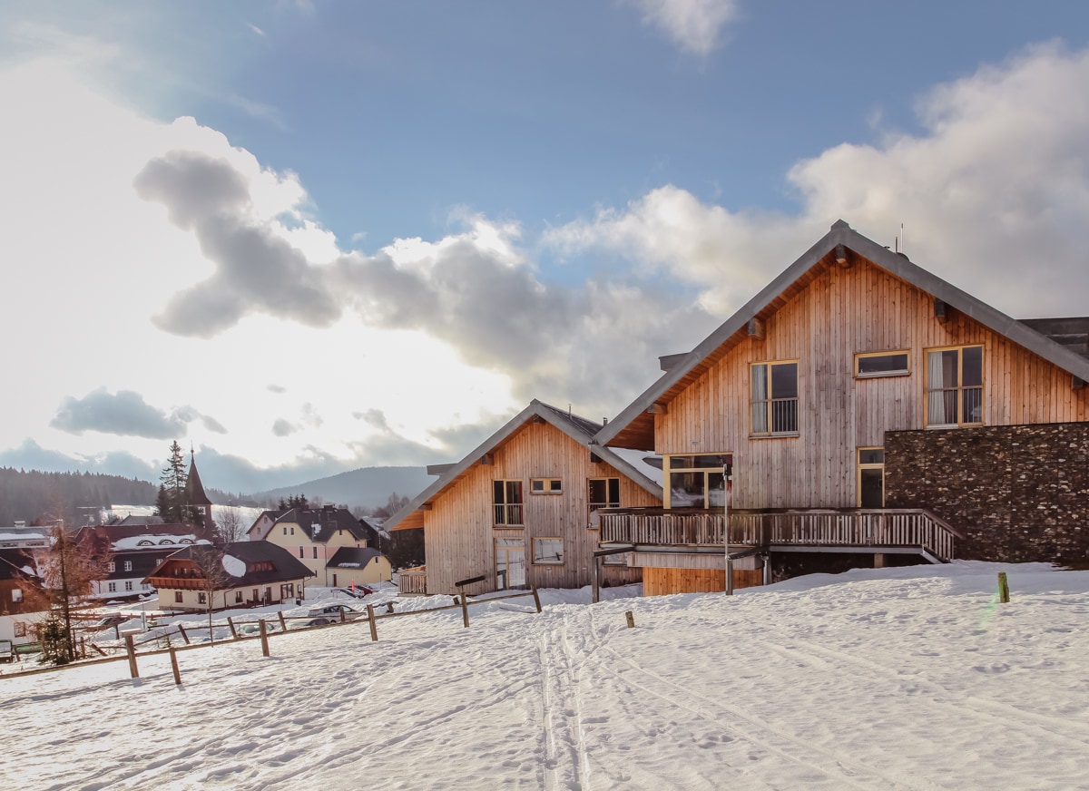 Two wooden buildings are situated in a snowy landscape, with a sloped roof design and large windows. In the background, hills and a residential area are visible under a partly cloudy sky, creating a serene winter atmosphere.