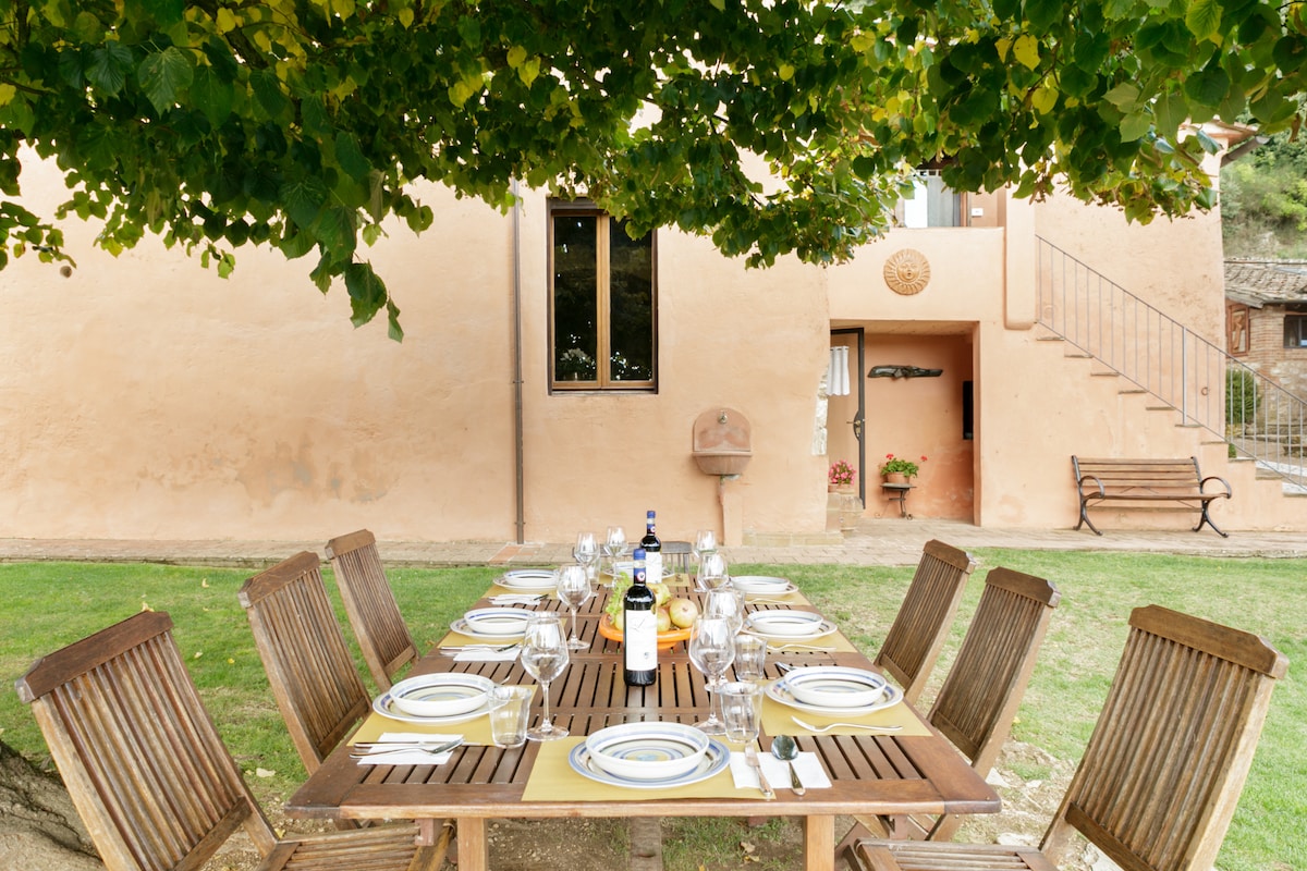 A spacious outdoor dining area is arranged under a leafy tree, featuring a large wooden table set with plates, glasses, and wine bottles. A stone wall and a bright facade of the villa provide a warm backdrop, while a staircase and a bench complete the inviting scene.