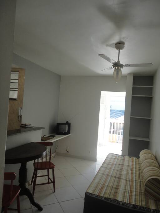 Foto da sala com sofá cama, ventilador de teto, prateleiras para acomodação de bagagens e utensílios pessoais, mesa redonda armável e cadeiras, rack com TV e utensílios de uso pessoal.
