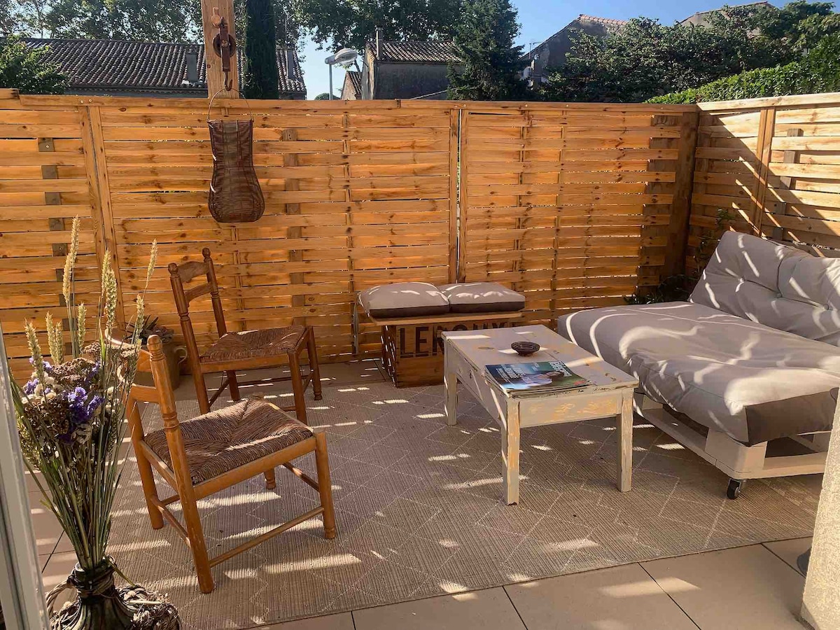 An outdoor space features a wooden fence that provides privacy. A comfortable lounge chair and two rustic chairs surround a small table. Natural light illuminates the area, highlighting the textured rug beneath. A cozy atmosphere is suggested, inviting relaxation or conversation.