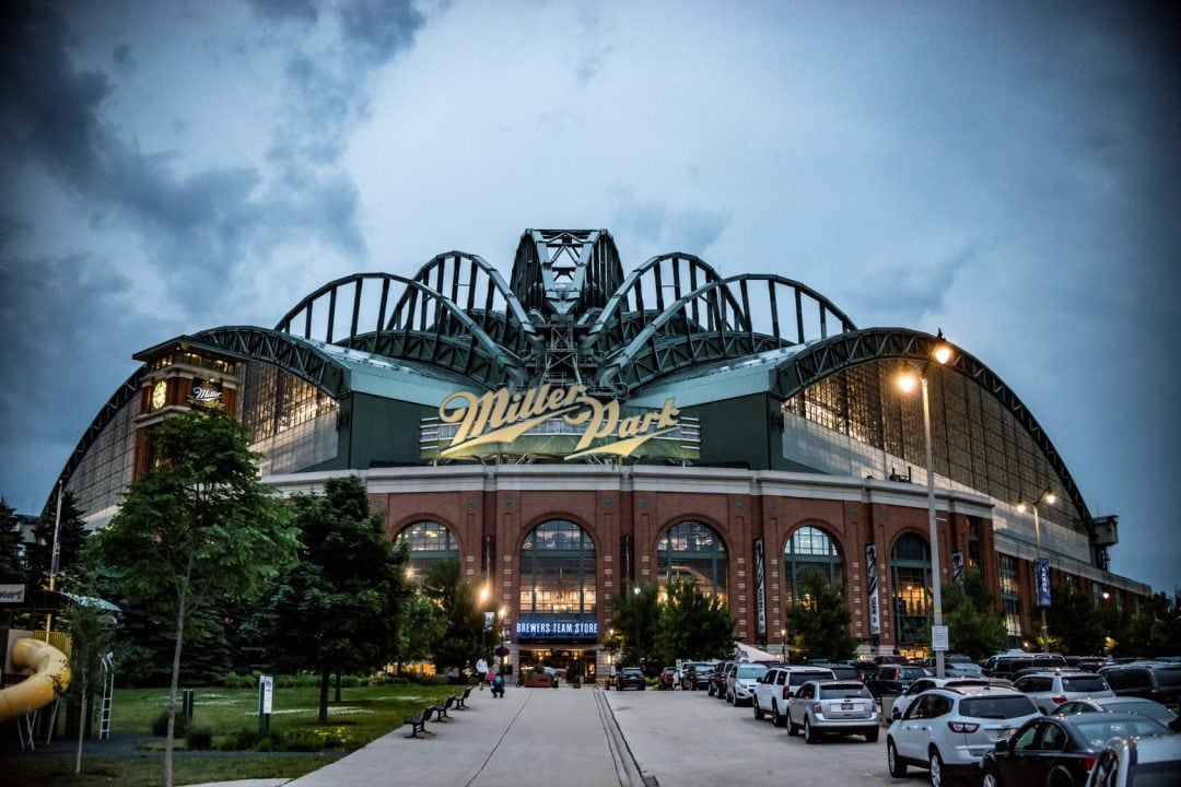 The exterior of a large stadium is showcased under a cloudy sky, featuring a distinctive arched roof structure. The entrance is illuminated, inviting visitors inside. Surrounding parking areas are visible, filled with vehicles, and landscaped greenery adds a natural touch to the scene.