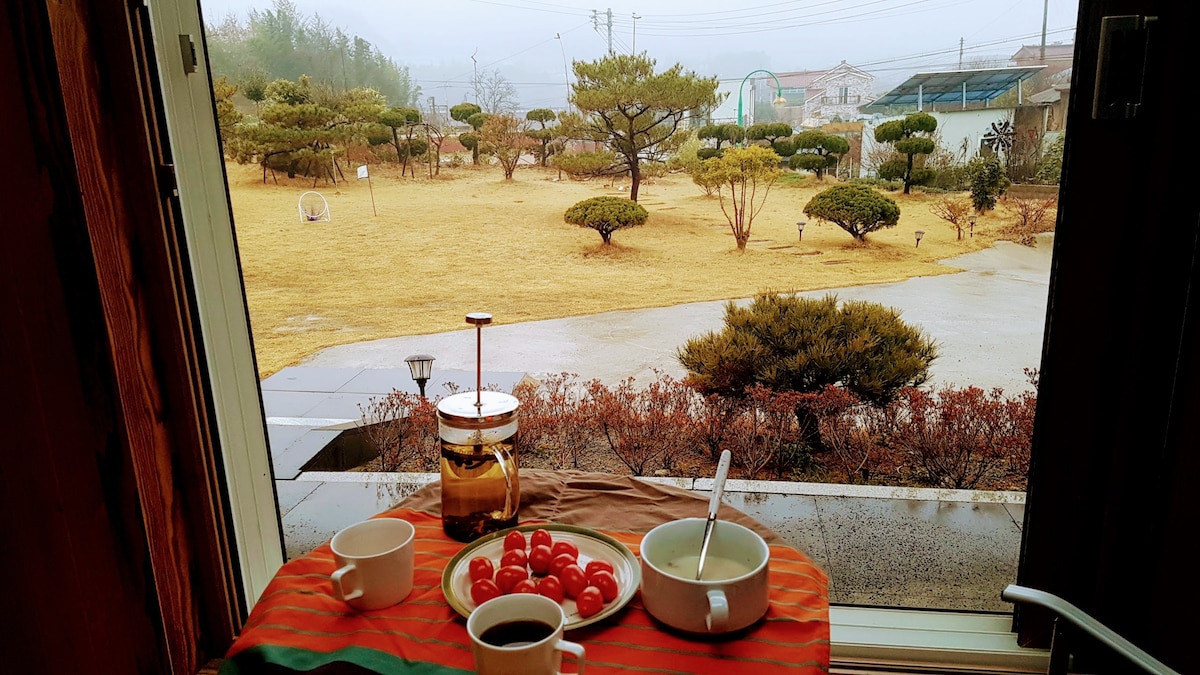 A window view showcases a spacious outdoor lawn, featuring manicured shrubs and trees. A cozy breakfast setting is arranged on a table with two cups, a French press, and a bowl of tomatoes. A serene atmosphere is created by the drizzling rain.