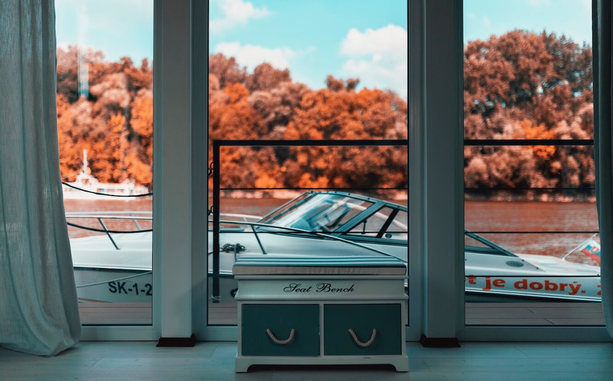 A view through large glass doors reveals a serene river scene, showing a boat docked nearby. Natural light filters in, creating a bright atmosphere, while a decorative chest with handles sits in the foreground, adding a touch of charm to the space.