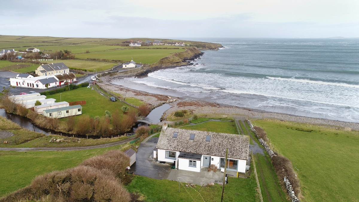 An aerial view reveals a charming beach front cottage set against the backdrop of the Atlantic Ocean. The surrounding landscape features a mix of green fields and a meandering stream. The foreground includes a parking area, while the beach stretches out along the shoreline.