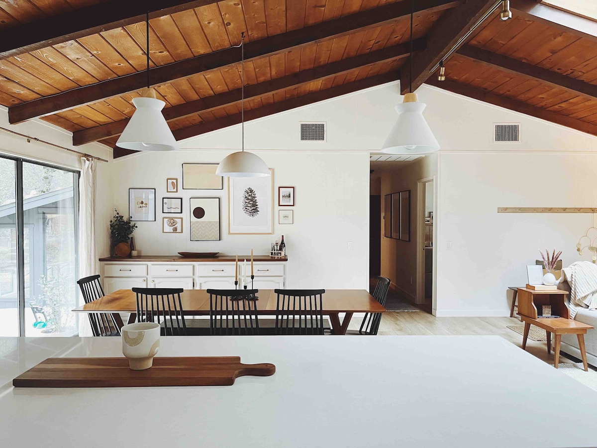 The open concept living area is highlighted by a high ceiling with exposed wooden beams. A large dining table is surrounded by black chairs, while a sideboard displays framed art. Natural light floods in through expansive glass doors, enhancing the inviting space.