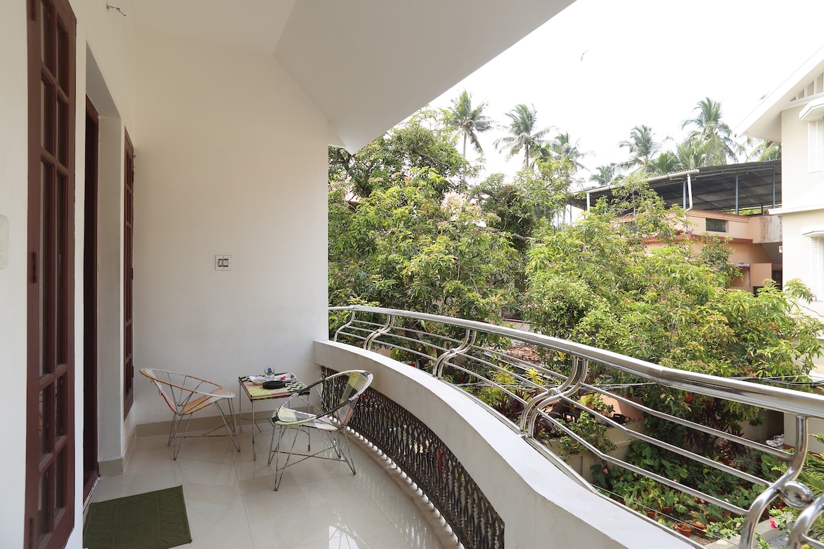 A balcony area features a small table and two chairs, surrounded by lush greenery and palm trees. The curved railing offers an unobstructed view of the vibrant foliage, creating a serene outdoor space for relaxation.