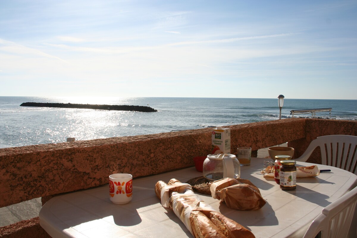A balcony table is set with fresh bread and breakfast items. The sea is visible in the background, shimmering under the sunlight. Expansive views of the beach and coastline create a serene setting for a morning meal.