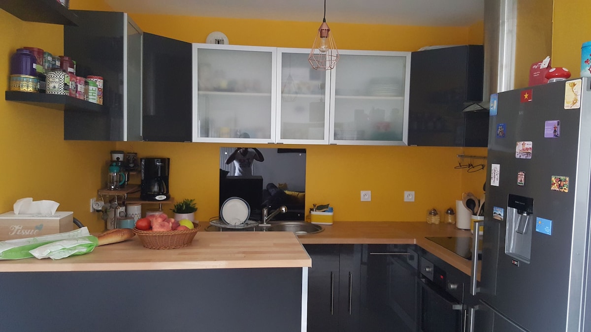 A modern kitchen features a bright yellow wall, complemented by dark cabinets and a wooden countertop. Open shelving displays various jars and kitchenware. A stainless steel refrigerator and appliances are present, with a small sink and coffee maker visible. A basket of fruit adds a touch of color.