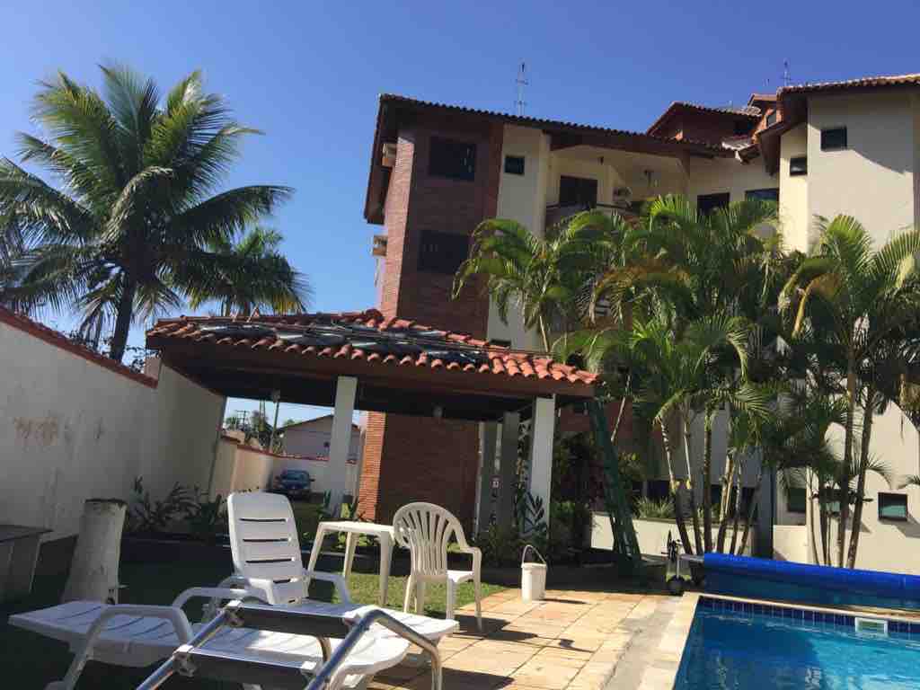 The image features a pool area with a tiled surface surrounded by palm trees. A shaded pavilion is supported by a slanted roof, with a few sun loungers positioned nearby. The building in the background is three stories tall with a terracotta roof.