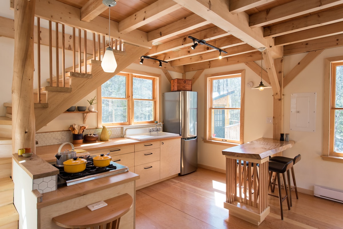 The open-concept kitchen showcases wooden beams and large windows that allow natural light to fill the space. A modern stove and stainless-steel refrigerator are complemented by wood cabinetry. A wooden dining table with tall stools is positioned near the counter, creating an inviting cooking and dining area.
