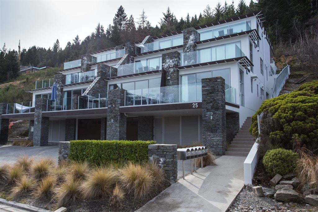 The apartment complex is presented with a modern architectural design, featuring multiple levels and large glass balconies. The building's exterior is complemented by dark stone features and neatly trimmed shrubs, enhancing the entrance pathway leading up to it.