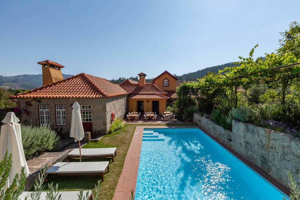 The outdoor area features a clear blue swimming pool surrounded by lounge chairs and well-maintained gardens. A house with a red-tiled roof is visible in the background, complemented by lush greenery and sunny skies.