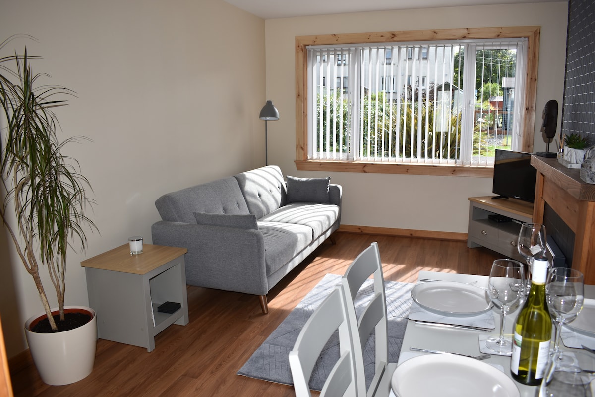 A bright living area features a grey sofa and a wooden side table, with a potted plant nearby. A large window with vertical blinds allows natural light to fill the space. A dining setup is visible, including a table set with plates and glasses.