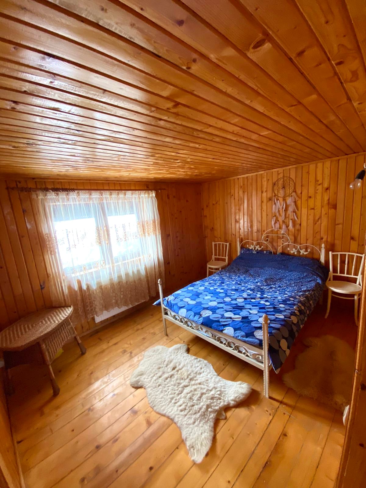 A warm wooden bedroom features a bed adorned with a blue patterned bedspread. Two small chairs are placed beside the bed, and a woven table sits in the corner. Natural light filters through the sheer curtains of a window, illuminating the wooden walls and floors.