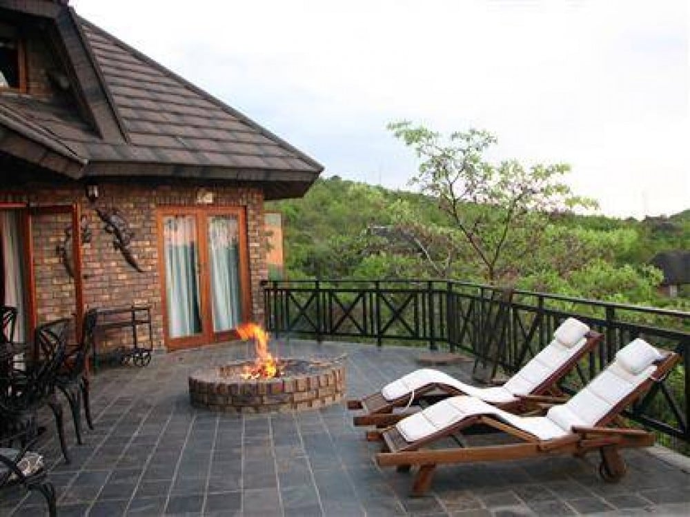 An outdoor deck features a circular stone fire pit with a small flame, surrounded by two white loungers for relaxation. The deck is bordered by a wooden railing, and a mix of greenery can be seen in the background, hinting at the natural beauty of the surrounding area.