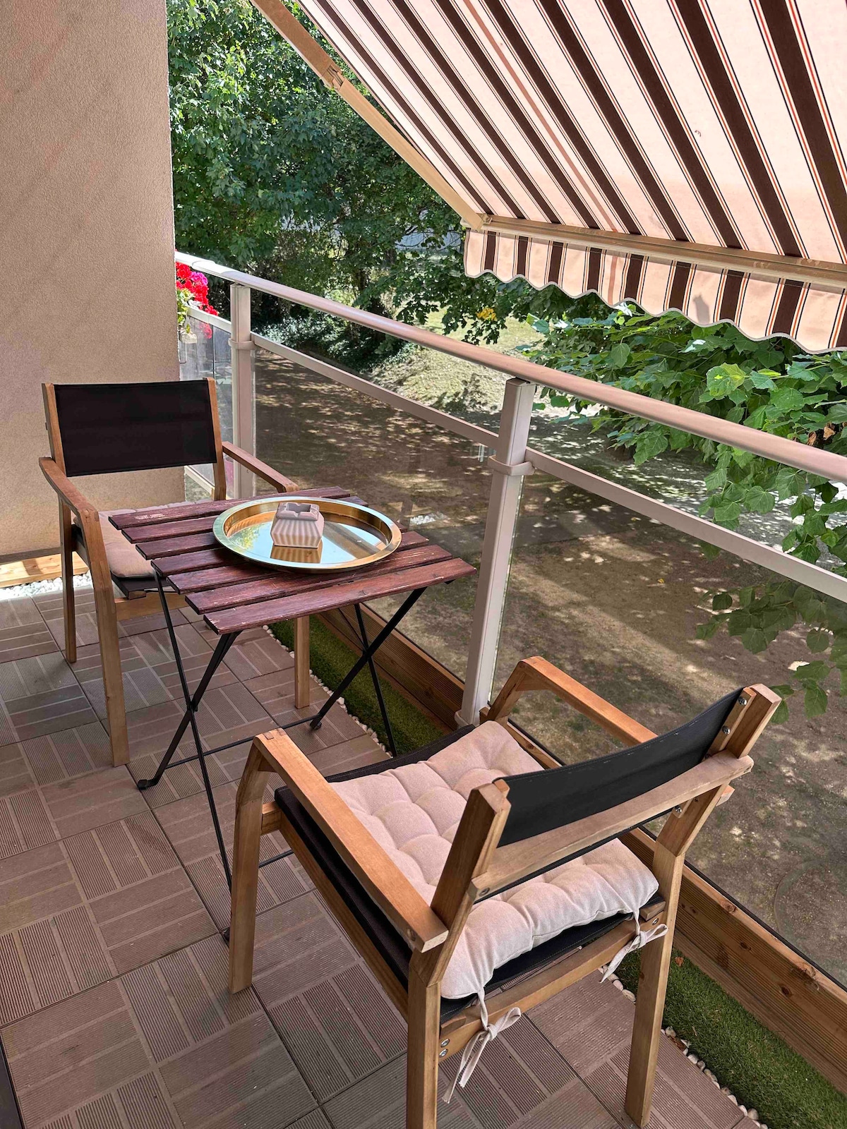 A small balcony features a wooden table and two chairs positioned under a retractable awning. A decorative tray rests on the table, surrounded by greenery from nearby plants, creating a serene outdoor space.