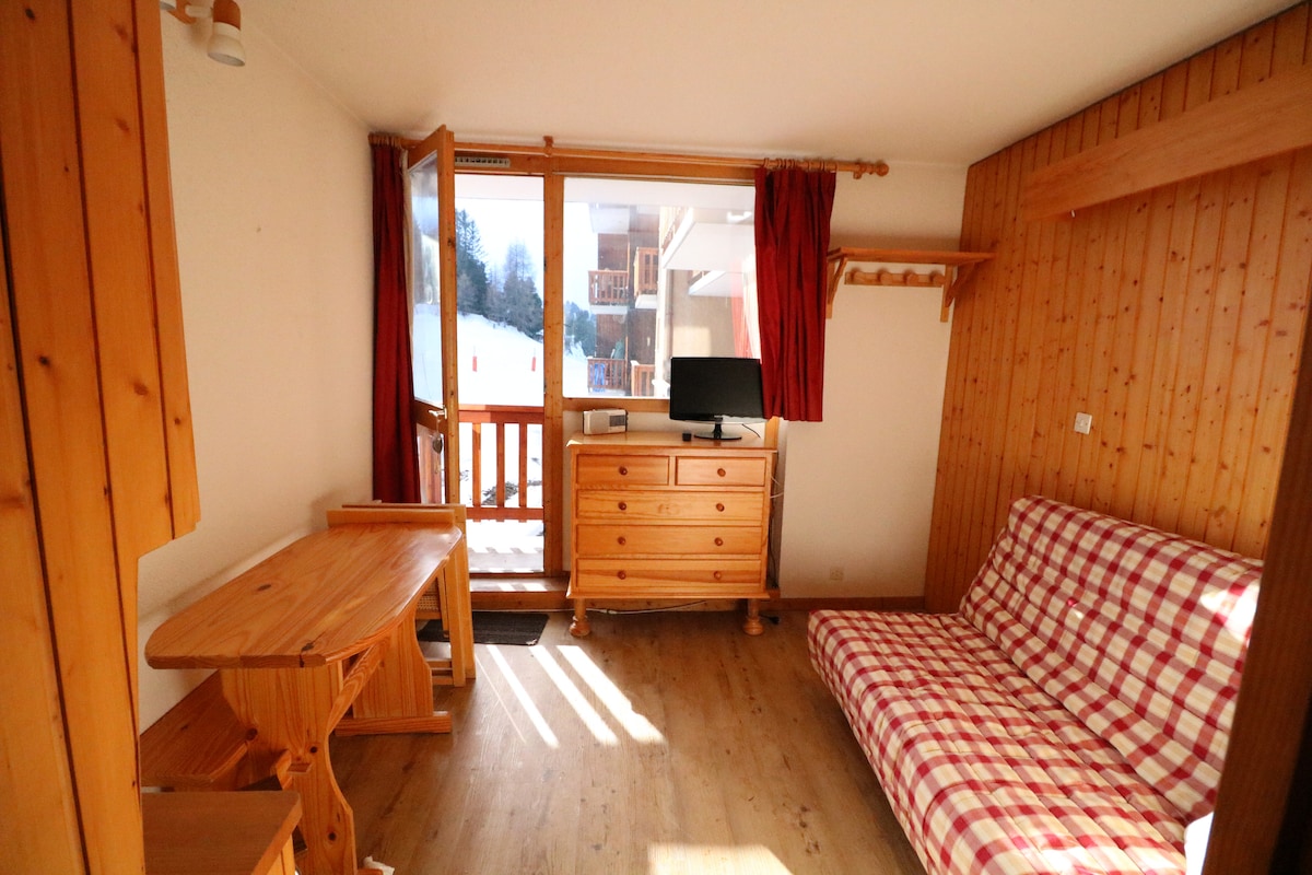 A well-lit interior features wooden accents and warm tones. A spacious seating area includes a fold-out couch with checkered fabric. A wooden dining table and chair set provide dining space, while a dresser and television are positioned against the wall. Large windows allow natural light to enter.