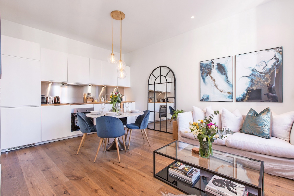 A modern living area features hardwood flooring and high ceilings. A marble dining table for four is set beside a fully fitted kitchen with sleek cabinetry. A comfortable sofa is accentuated by decorative pillows, and a glass coffee table displays books and fresh flowers.