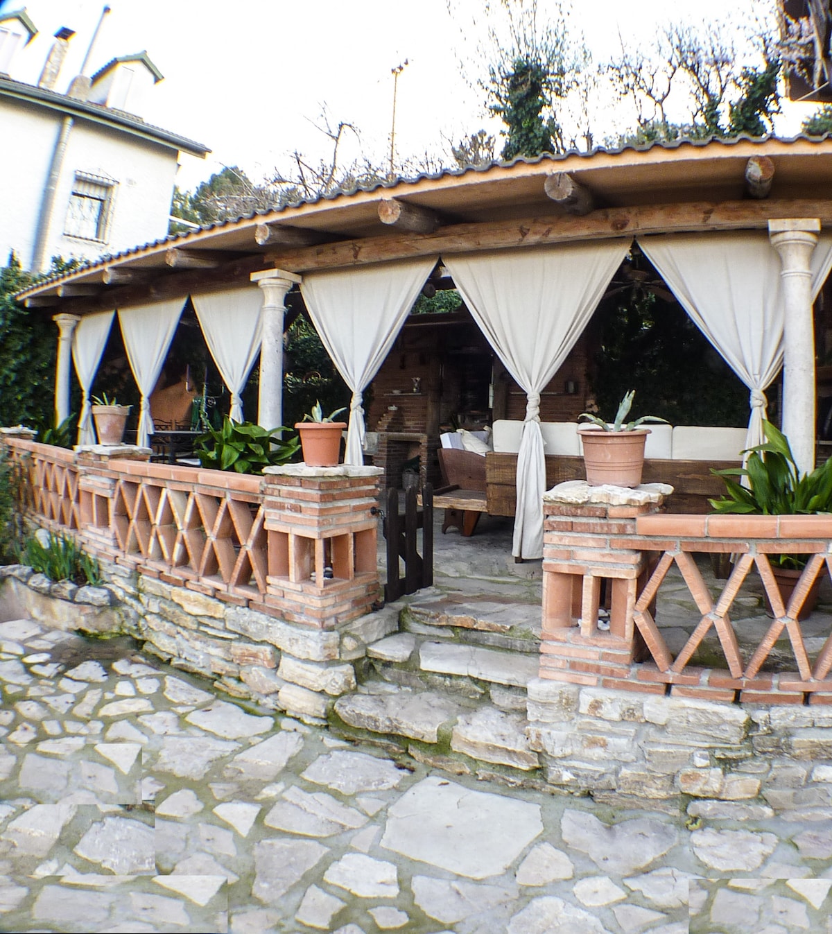 A covered porch area is framed by draped curtains and features comfortable seating. Stone pathways lead to the entrance, surrounded by potted plants. The rustic wood elements of the structure create a welcoming atmosphere, inviting relaxation in a shaded outdoor space.