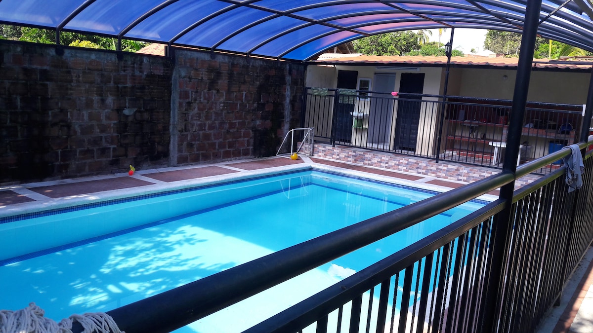 A private swimming pool is enclosed under a transparent canopy, showcasing clear blue water. Surrounding the pool, a black metal fence provides safety and boundary. The area features a tiled deck and is accompanied by a shaded lounging space for relaxation.