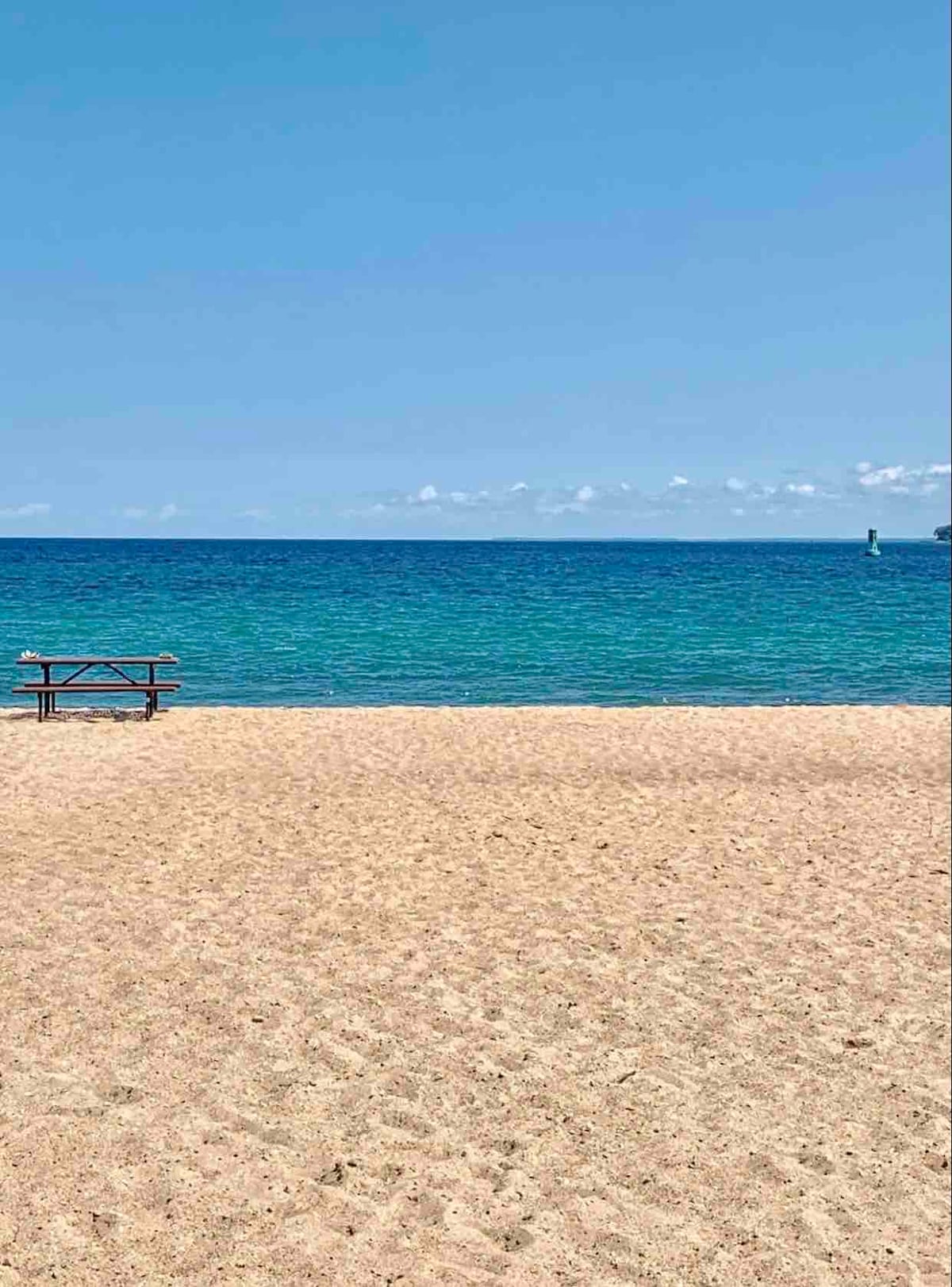 A sandy beach stretches out, meeting a calm blue lake under a clear sky. A wooden picnic table is positioned on the sand, providing a space for relaxation and enjoyment of the scenic view. The horizon blends harmoniously with the water.