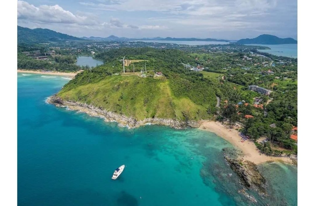 An aerial view of a lush green hill overlooking clear turquoise waters. A small boat is anchored in the calm sea, with sandy beaches visible along the shoreline. The landscape includes distant hills and clusters of vegetation, offering a serene coastal scenery.