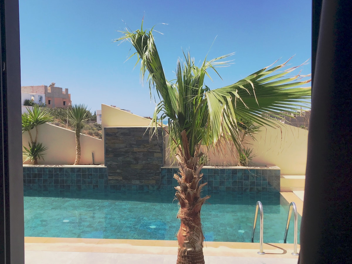 A view of a serene swimming pool is framed by a vibrant palm tree. The pool features clear blue water and is surrounded by a stone wall, with a bright sky visible in the background.