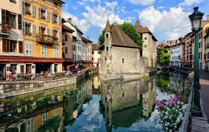 A peaceful scene along a canal reflects historic buildings with colorful facades and charming architecture. Flowering plants line the walkway beside the water, while clouds drift overhead, enhancing the ambiance of the serene waterfront in the town center.