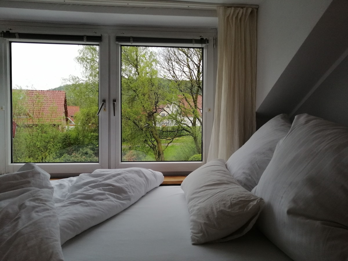 Natural light fills the space through large windows, framing views of green trees and rooftops. A spacious bed is adorned with soft pillows and white linens, creating a serene and welcoming environment.