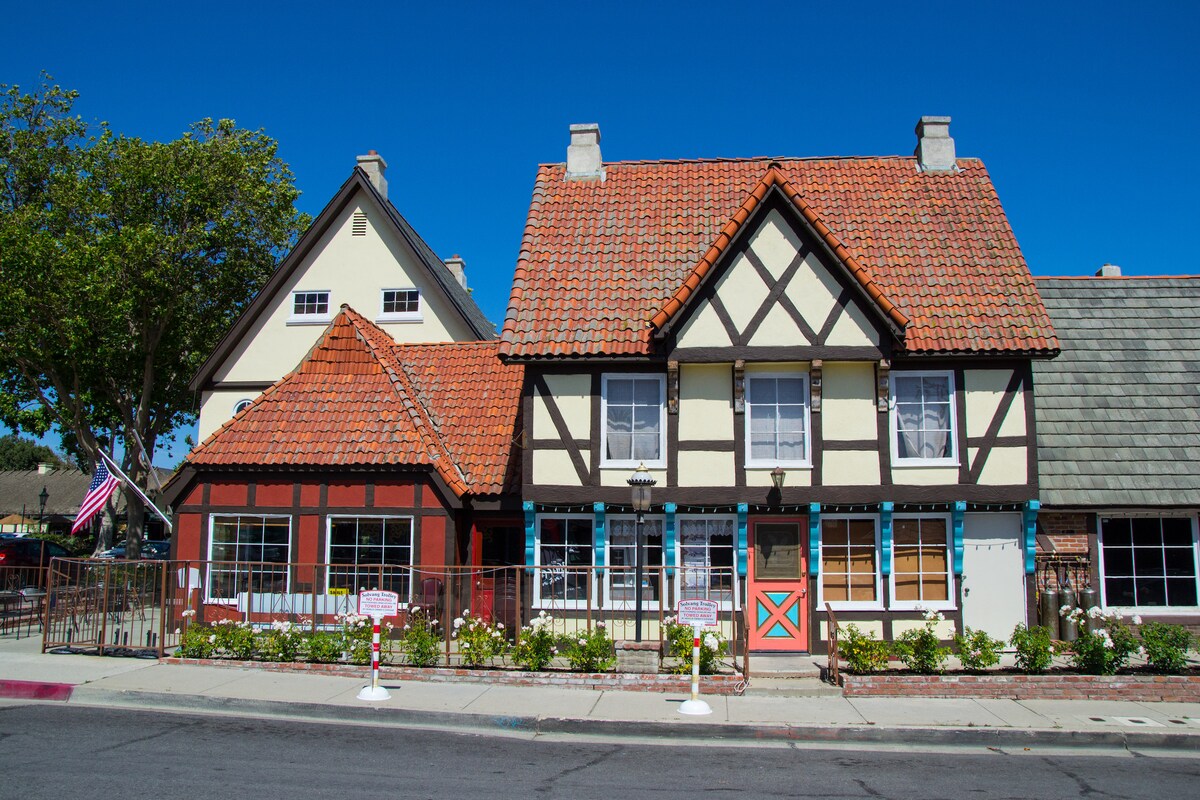 A charming architectural facade is shown, featuring a combination of red and cream colors with dark timber accents. The roofs are covered with terracotta tiles. Neatly arranged flowers line the front, and a flag signifies a welcoming atmosphere. A clear blue sky provides a bright backdrop.