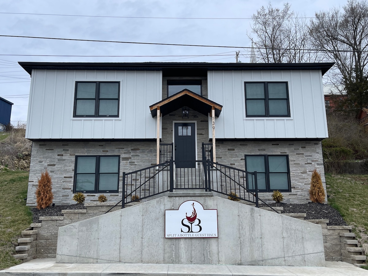 A modern split-level guest house is displayed, featuring a combination of gray stone and white panels. A welcoming entrance is flanked by neatly landscaped areas and decorative shrubs. The property is situated on a gentle slope with a sign at the base indicating the guest house's name.