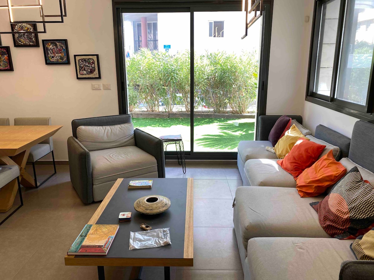 A comfortable living area features a large gray sofa adorned with colorful cushions, alongside a cozy armchair. A wooden coffee table with decorative items is centered in the space. Bright natural light fills the room, highlighting the greenery visible through the sliding glass doors.