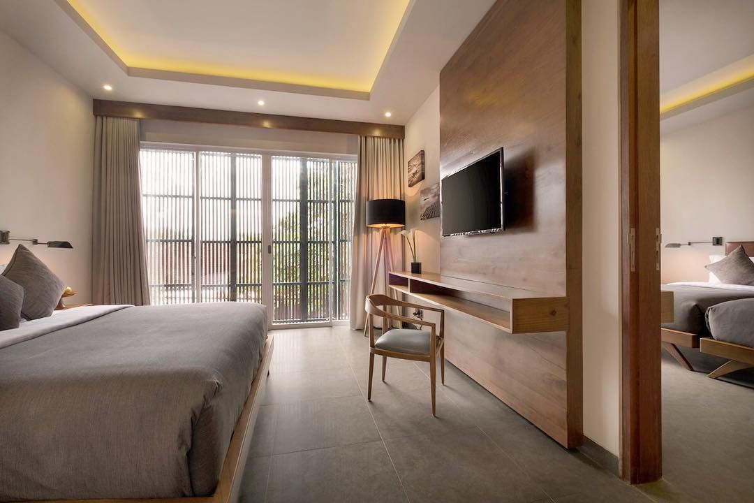 A spacious room features a large bed with soft linens, complemented by modern wooden accents. Large glass doors allow natural light to fill the space, leading to an outdoor view. A flat-screen TV is mounted on the wall, and a stylish desk chair is positioned near a compact desk.
