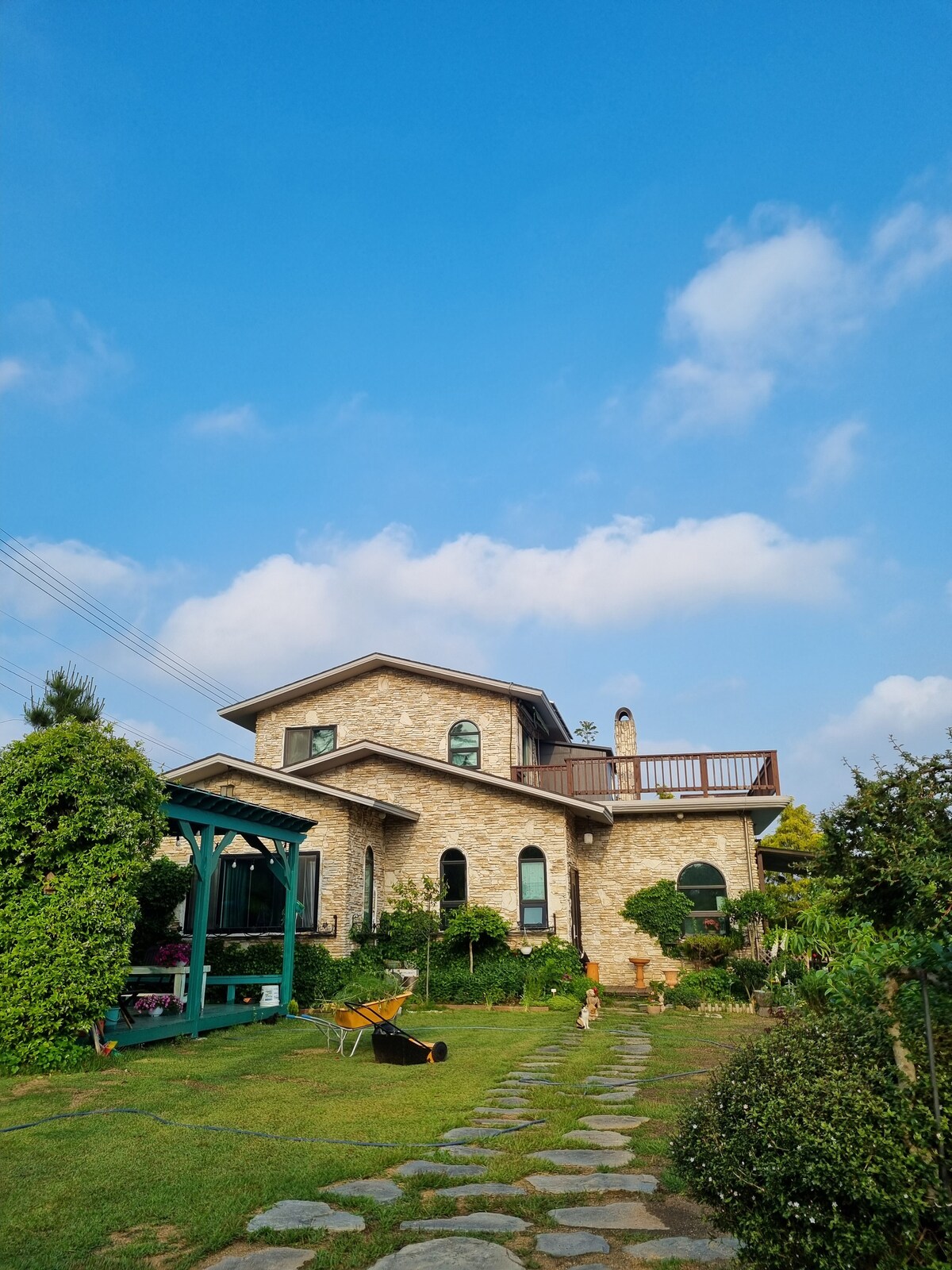 A two-story house with a stone facade is surrounded by greenery and a well-maintained lawn. A stone path leads to the entrance, with large windows allowing natural light. The balcony is visible on the upper level, and a wooden gazebo is situated nearby.