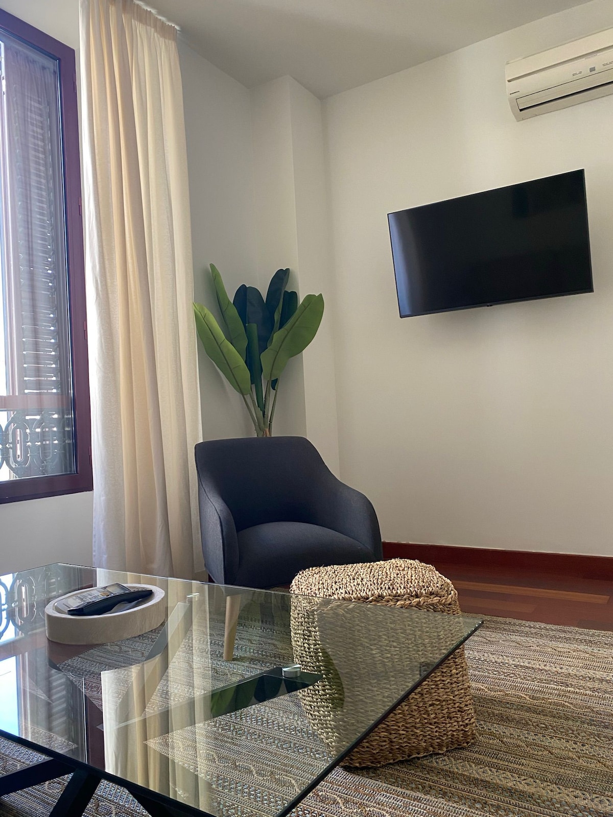 A comfortable seating area is featured, consisting of a modern chair and a woven ottoman. A flat-screen television is mounted on the wall. Natural light is invited through large windows with sheer curtains, creating a bright and airy atmosphere.