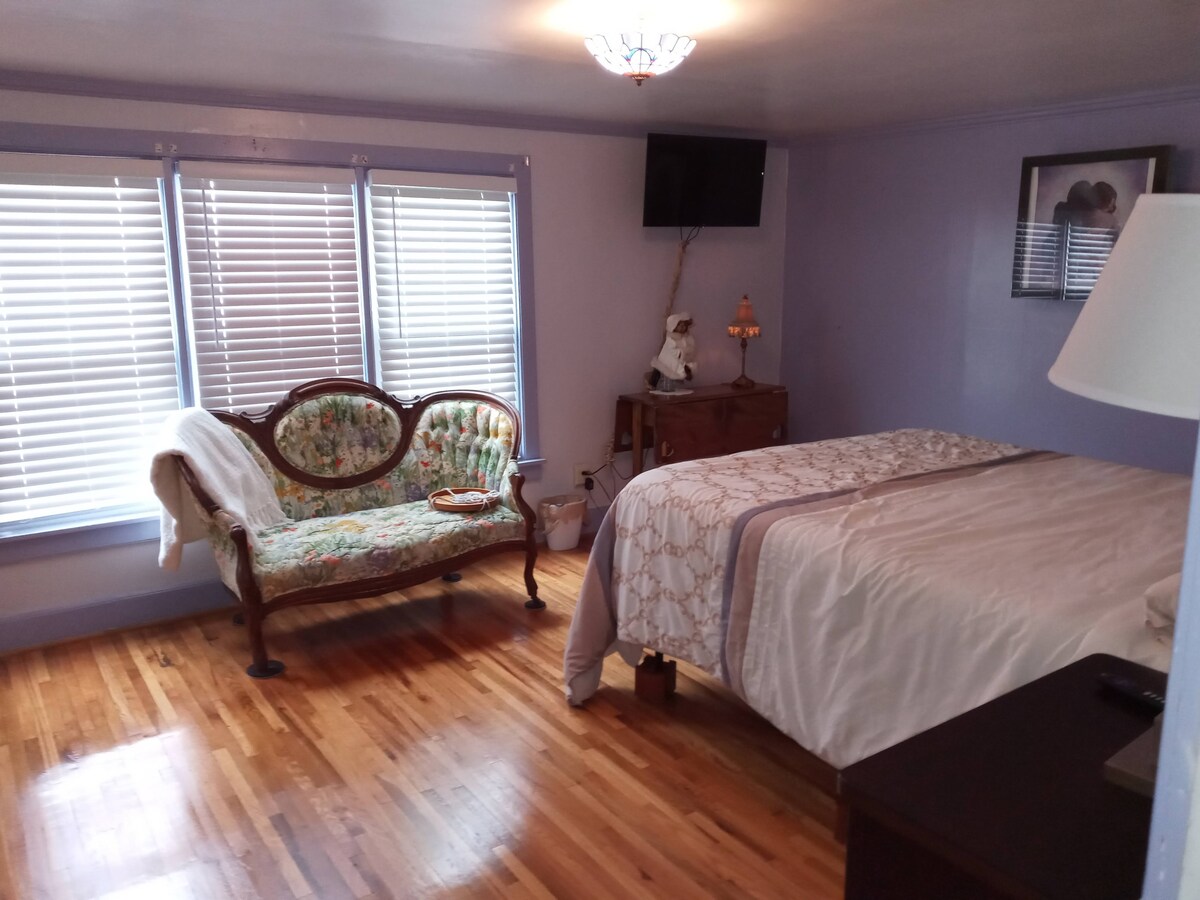 An upstairs bedroom features a spacious bed with decorative bedding, paired with a vintage-style settee. Natural light enters through three windows, which are dressed with blinds. A television is mounted on the wall, with wooden flooring complementing the room's tranquil purple walls.