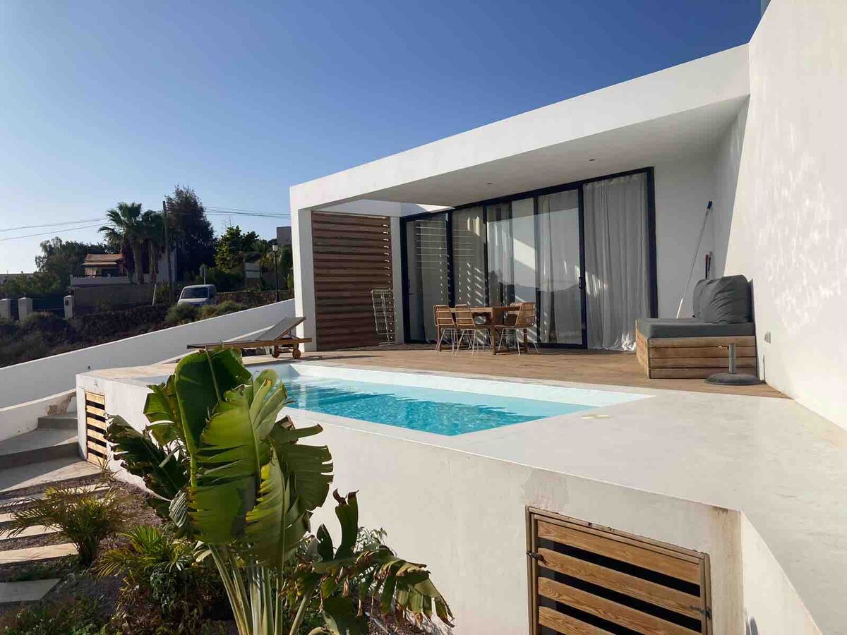 A modern house with a private swimming pool is showcased in this image. Sliding glass doors open to a spacious terrace featuring a wooden dining table and a comfortable lounge area. Lush greenery surrounds the pool, and the clear sky enhances the inviting outdoor space.