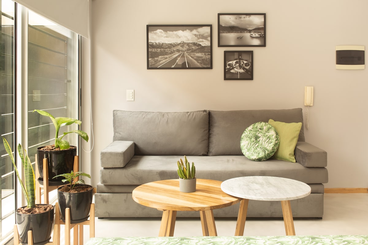 A modern living area features a gray sofa adorned with a green patterned cushion. Two coffee tables, one with a marble top, are set in front of the sofa. Potted plants are positioned beside the window, enhancing the space with vibrant greenery.