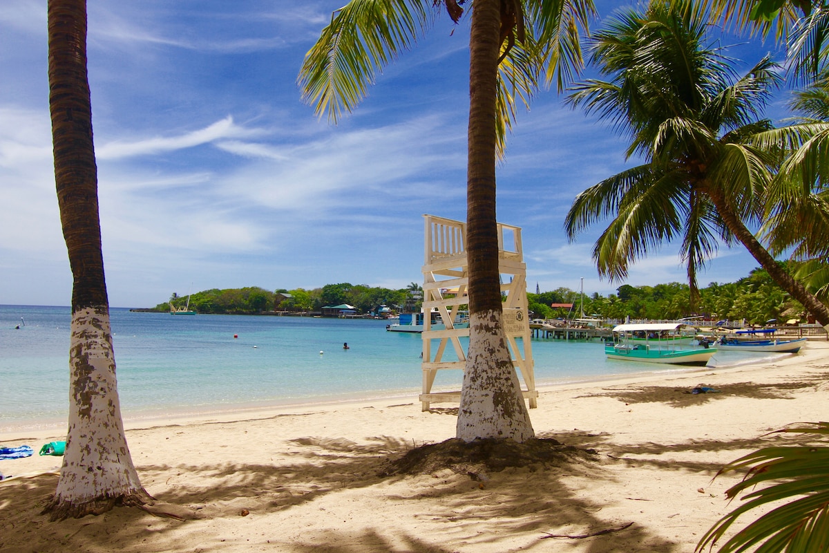 A serene beach scene is depicted with gentle waves lapping at the shore. Two palm trees frame the image alongside a lifeguard chair. A clear blue sky and distant boats add to the tranquil atmosphere, inviting relaxation near the water's edge.