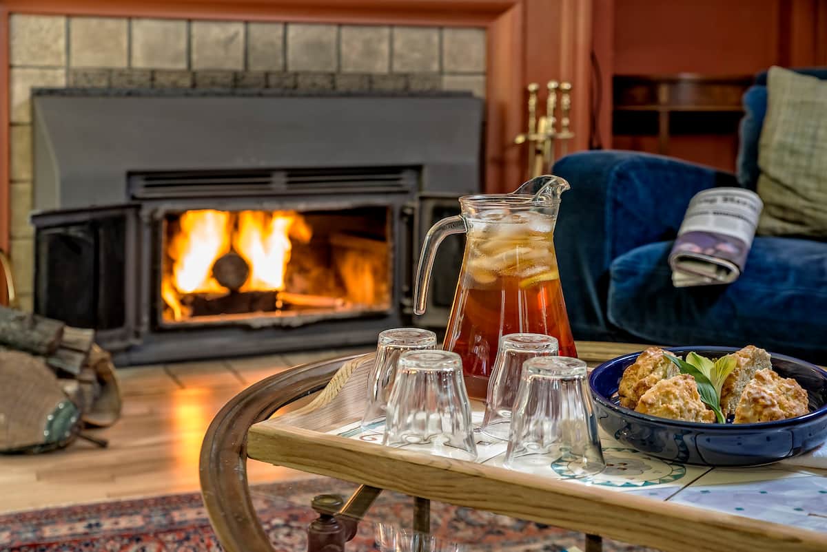 A warm living area features a fireplace with a soft glow, illuminating a stylish coffee table set with a pitcher of iced tea and several glasses. A bowl filled with baked goods is arranged nearby, enhancing the inviting atmosphere.
