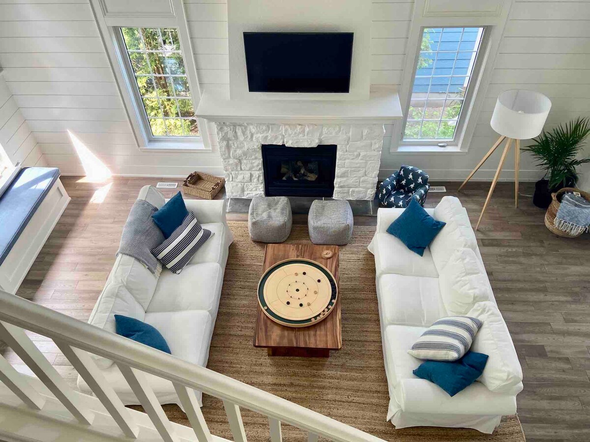 An airy living space features two white sofas adorned with blue and gray pillows, arranged around a wooden coffee table. A stone fireplace serves as the focal point, while large windows allow natural light to illuminate the room. A decorative floor lamp and indoor plants add to the ambiance.