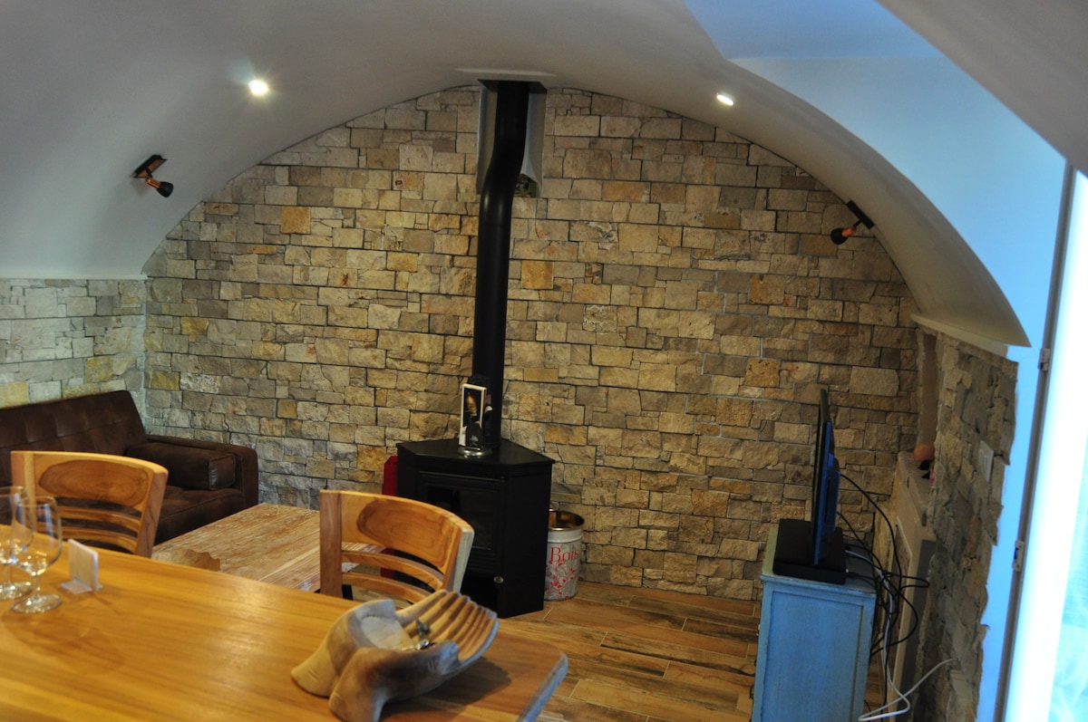 An inviting living space features a curved ceiling and stone accent wall, complemented by a cozy wood stove. A rustic dining table is positioned nearby, with wooden chairs arranged thoughtfully. A television stands on a small blue cabinet, enhancing the area’s functionality.