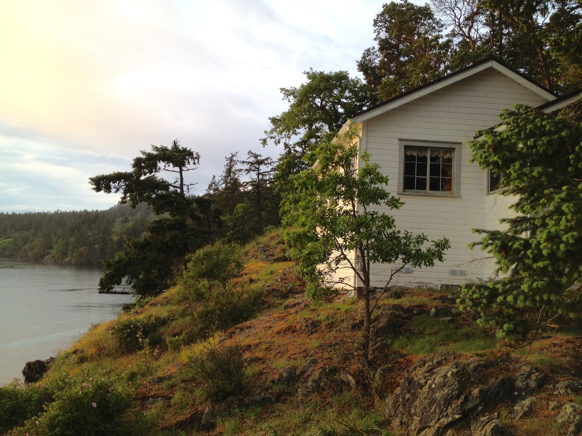 A vintage cabin is nestled on a rocky hill, surrounded by lush greenery. The building features large windows, allowing natural light to fill the interior. The nearby Salish Sea is visible, reflecting the soft colors of the sky during twilight.